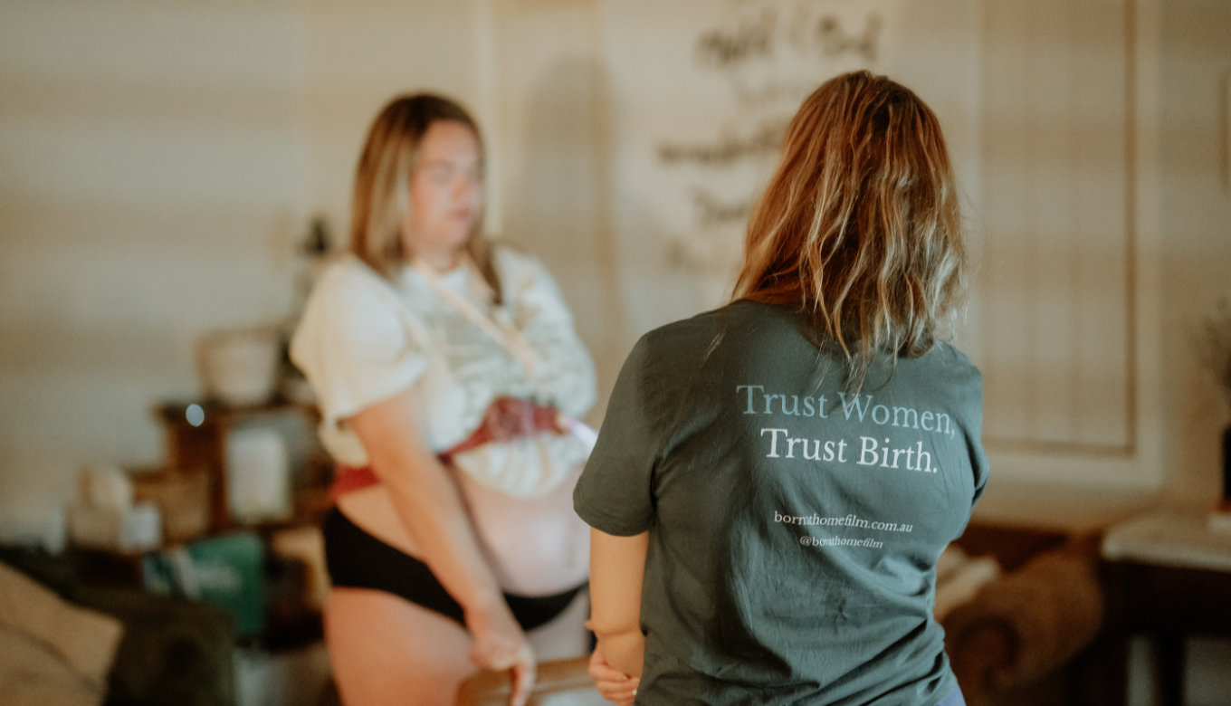 Alyssa Booth wearing a shirt which says 'Trust Women, Trust Birth', a woman is labouring out of focus in the background