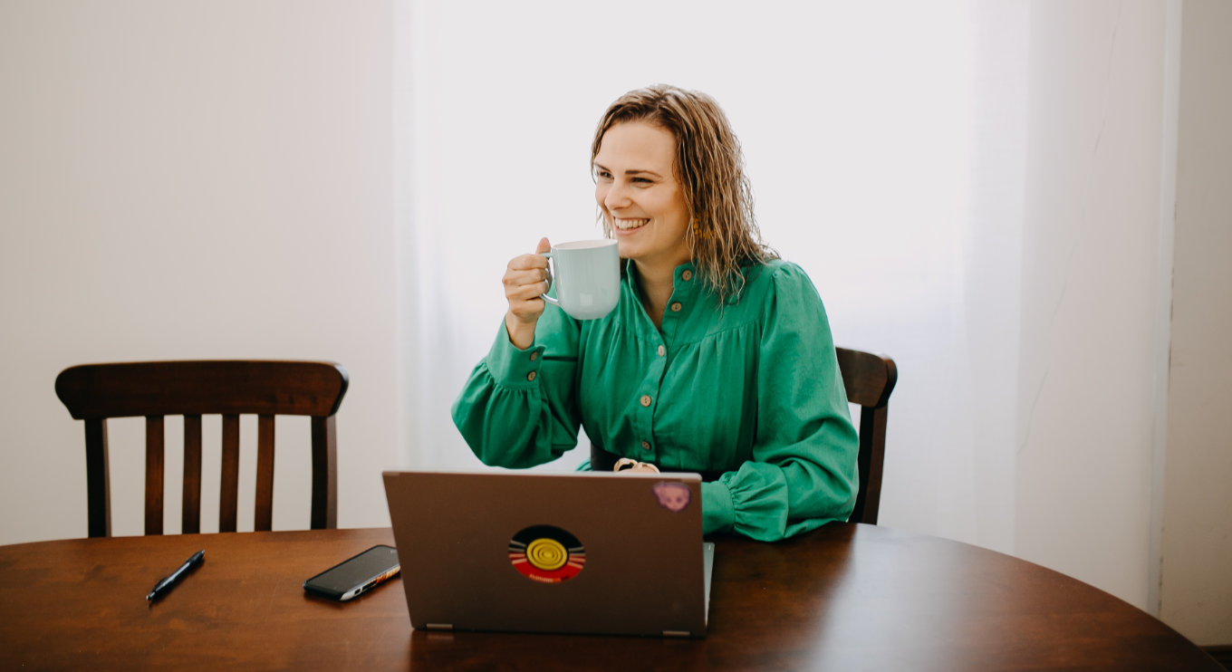 Alyssa Booth wearing a green dress sitting at a table with a laptop and holding a mug of tea