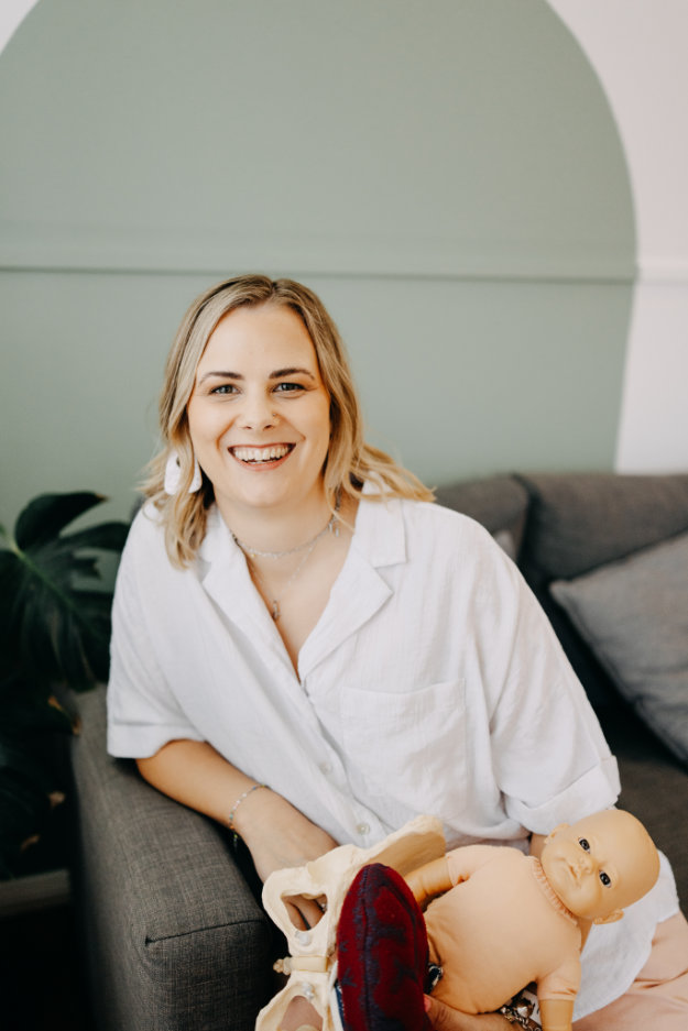 Photo of Alyssa Booth smiling on a sofa while holding a pelvis model and baby doll for birth education